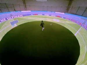 Player trains inside a SoccerBot360 arena, striking a ball on indoor turf surrounded by curved rebound boards.