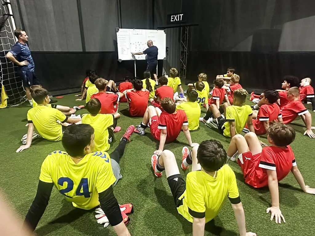 A full squad of youth soccer players seated on the indoor turf for a tactical session at Soccer Field Academy in Columbus, Ohio