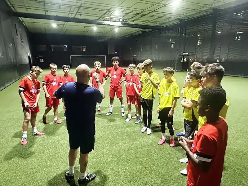 Youth soccer players gathered around their coach during an evening session on the indoor turf at Soccer Field Academy in Columbus, Ohio